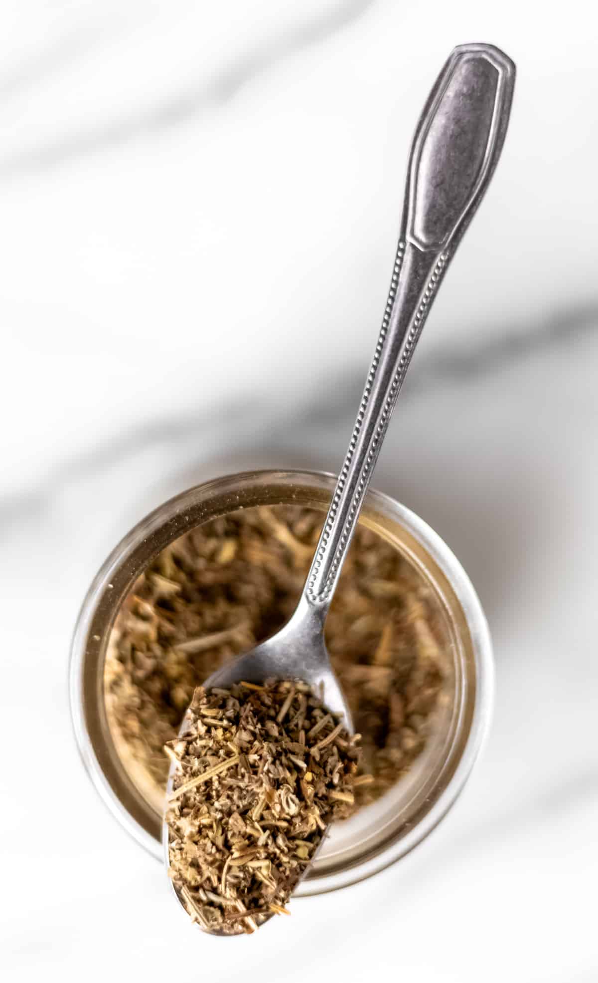 A spoonful of Italian seasoning sitting on top of a glass spice jar.
