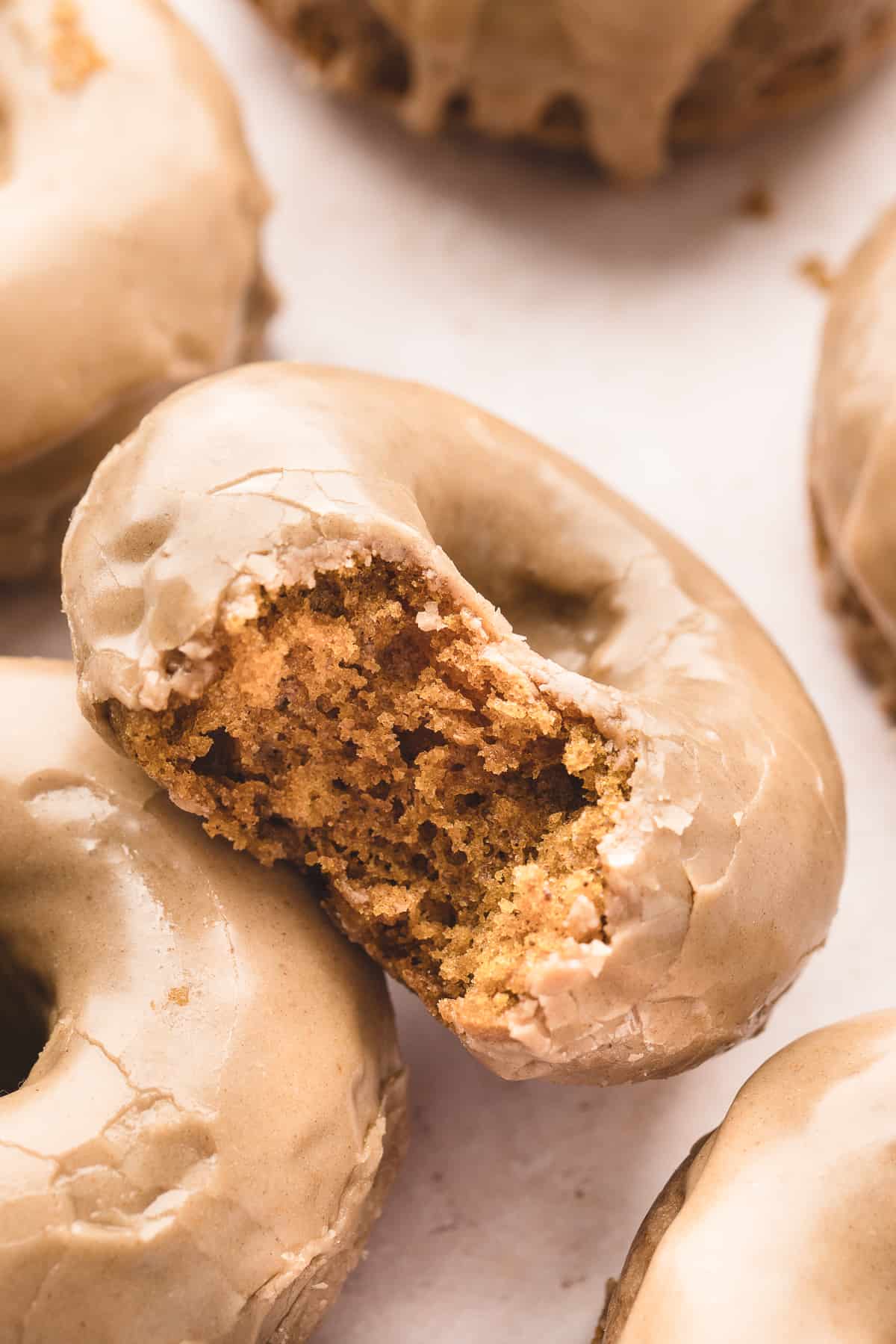 A pumpkin donut with maple glaze tilted up with a bite taken out to show the inside with other donuts around it.