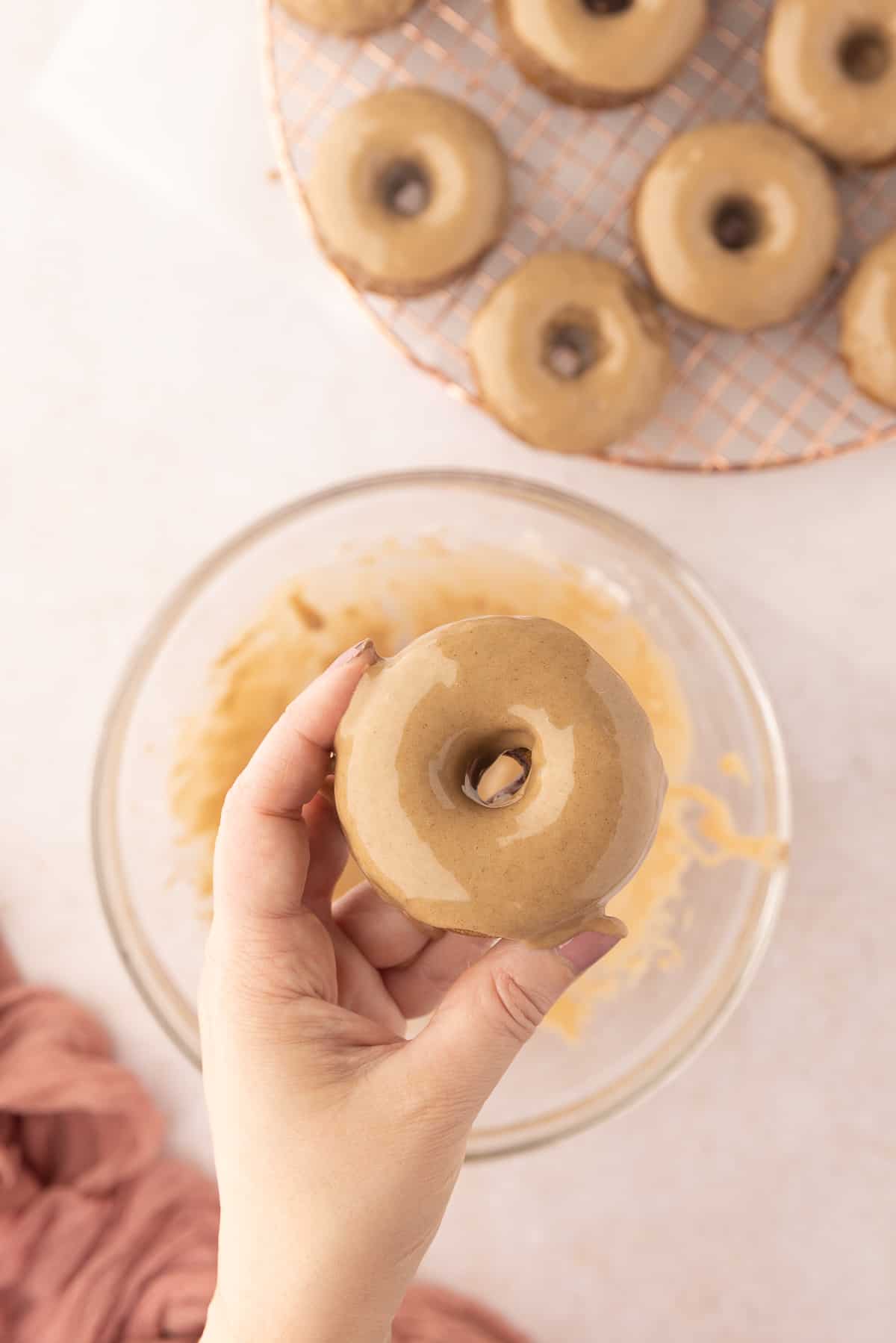 A donut being held up that has just been dipped into a bowl of cinnamon maple glaze under it, with a cooling rack of more donuts in the background.