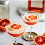 Two glasses of blood orange amaretto sour with oranges and the bottle around them.