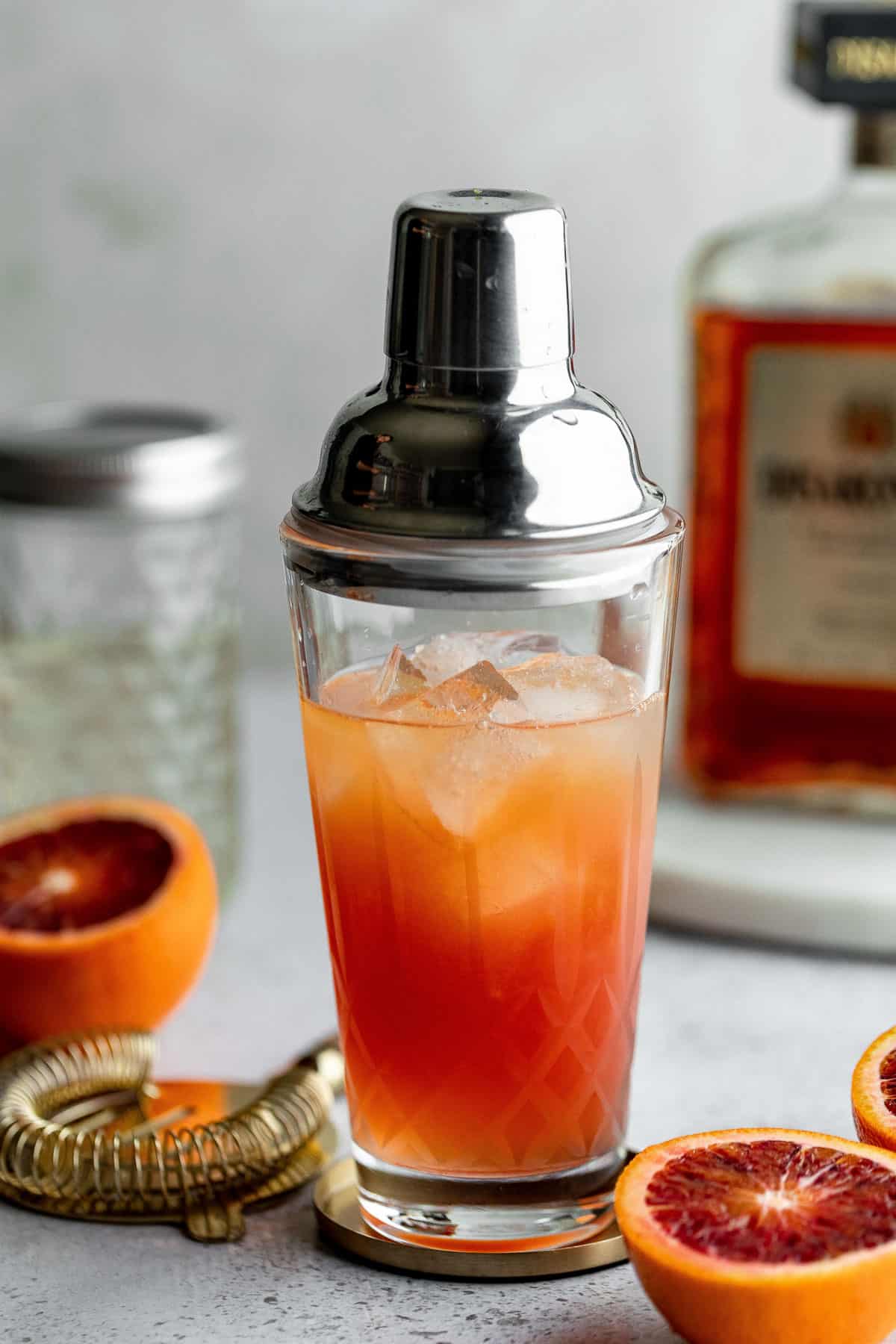 Blood orange amaretto sour in a cocktail shaker with bottles and blood oranges.