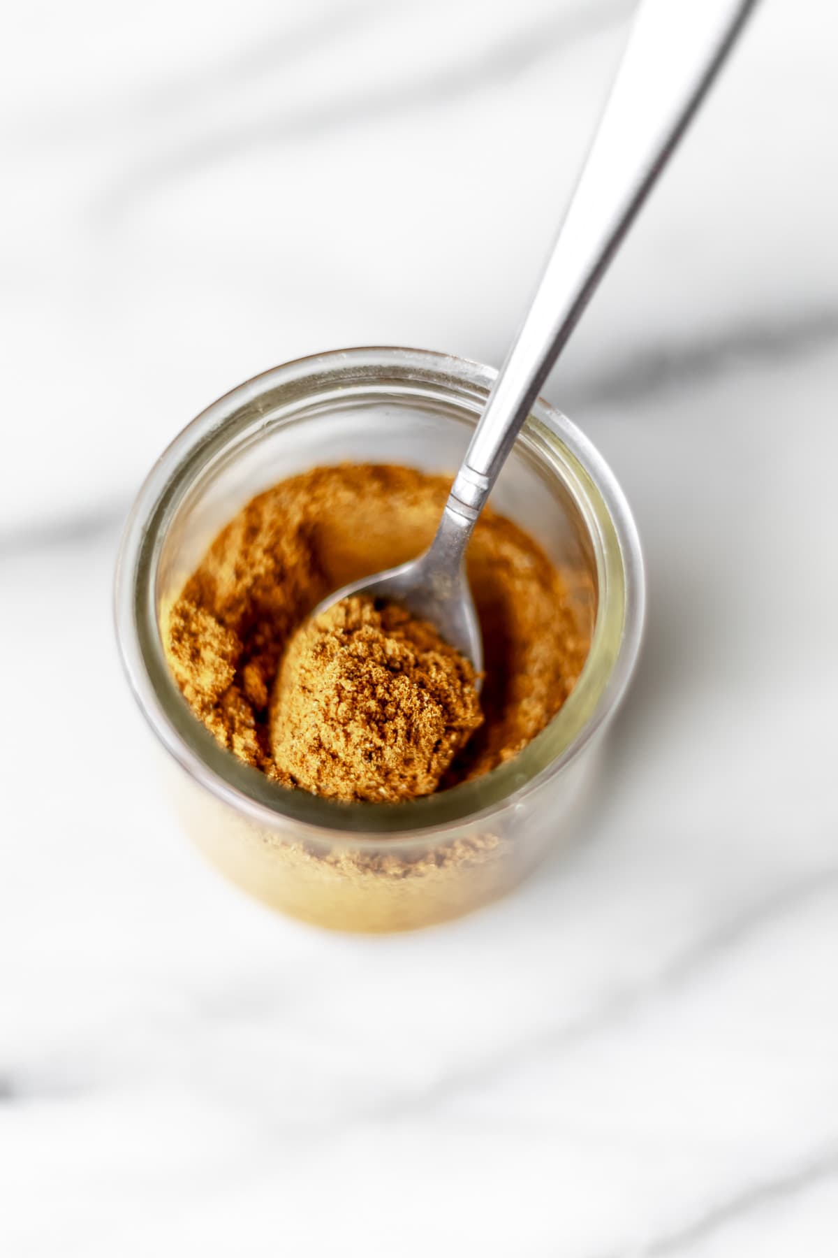 A glass jar of curry powder with a small spoon in it over a marble background.