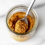 Close up of a small glass jar of curry powder with a small spoon in it.