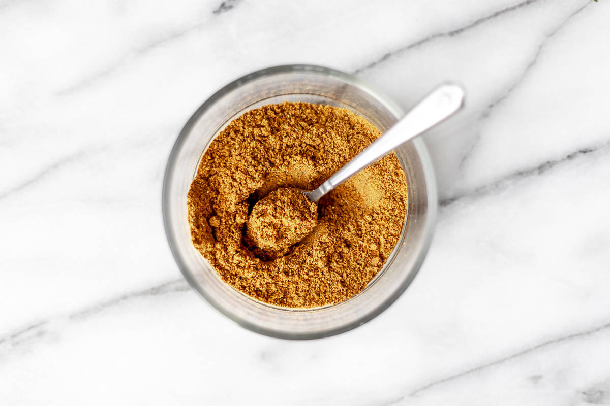 Homemade curry powder in a small glass bowl with a spoon in it over a marble background.