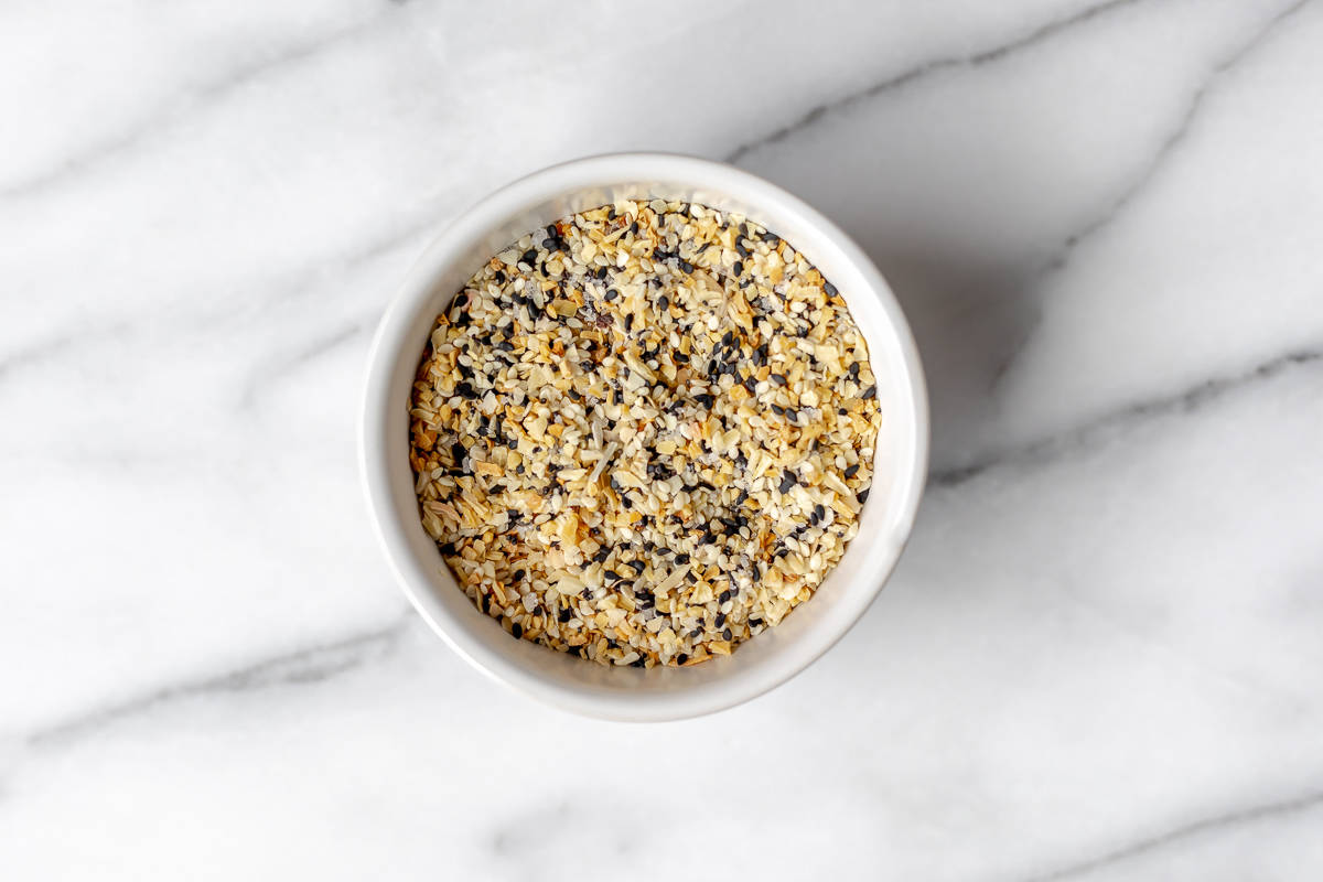 Overhead of everything bagel seasoning in a small white bowl over a marble background.