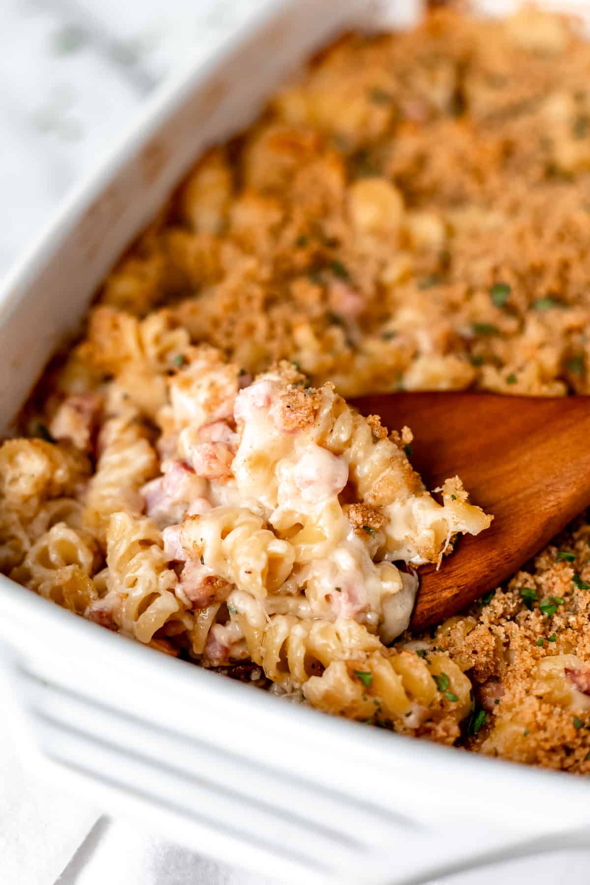 A ham and cheese pasta casserole with a wood server scooping some up in the baking dish.