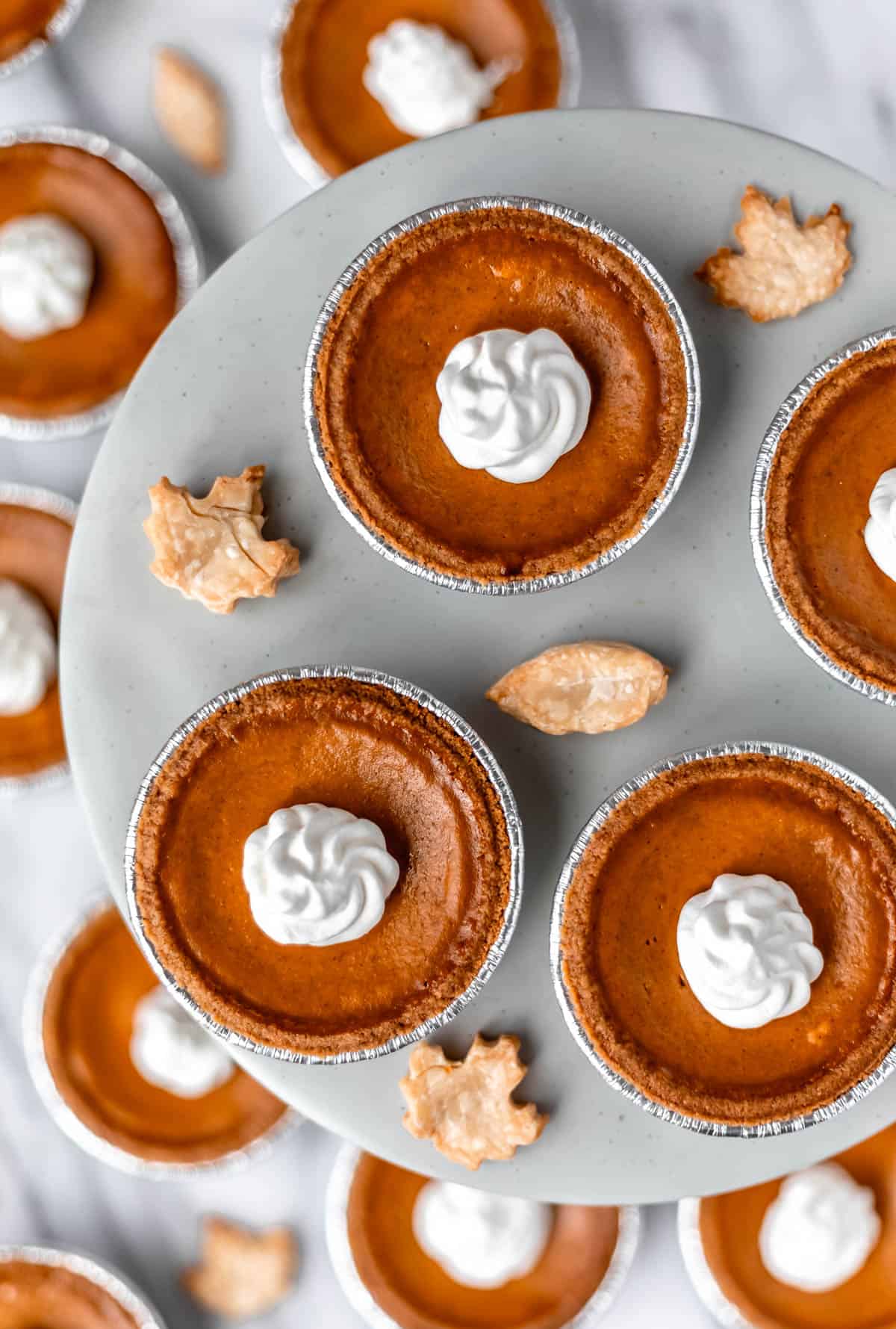 Overhead of mini pumpkin pies on a gray pie stand and around it with pie crust leaves around them.