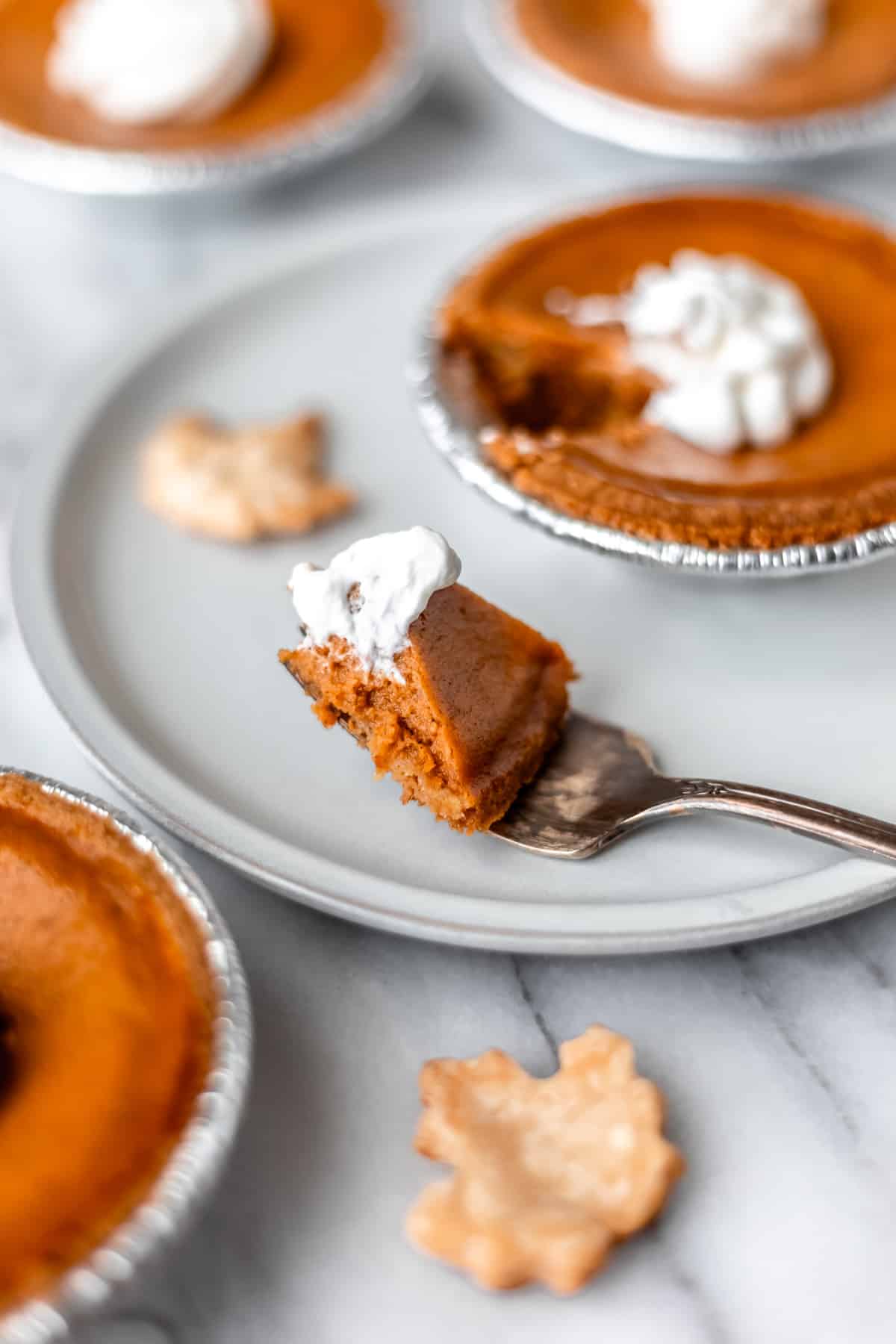 A bite of pumpkin pie and a mini pie on a plate with other mini pies and pie crust leaves around it.