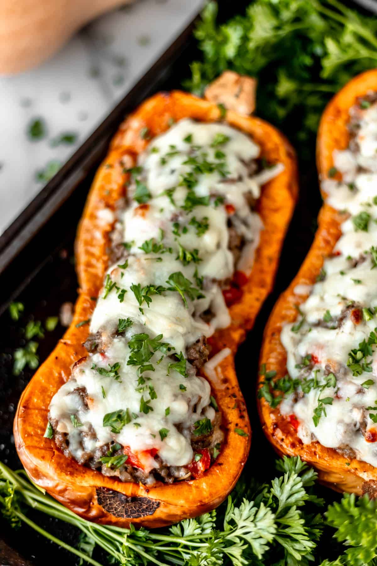 Two stuffed butternut squash halves on a baking sheet with parsley around it and a butternut squash in the background.