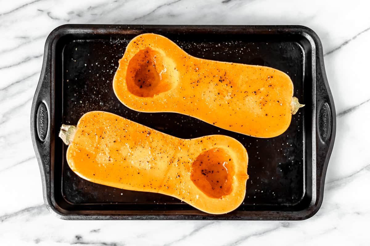 Two butternut squash halves drizzled with olive oil and seasoned with salt and pepper on a baking sheet.