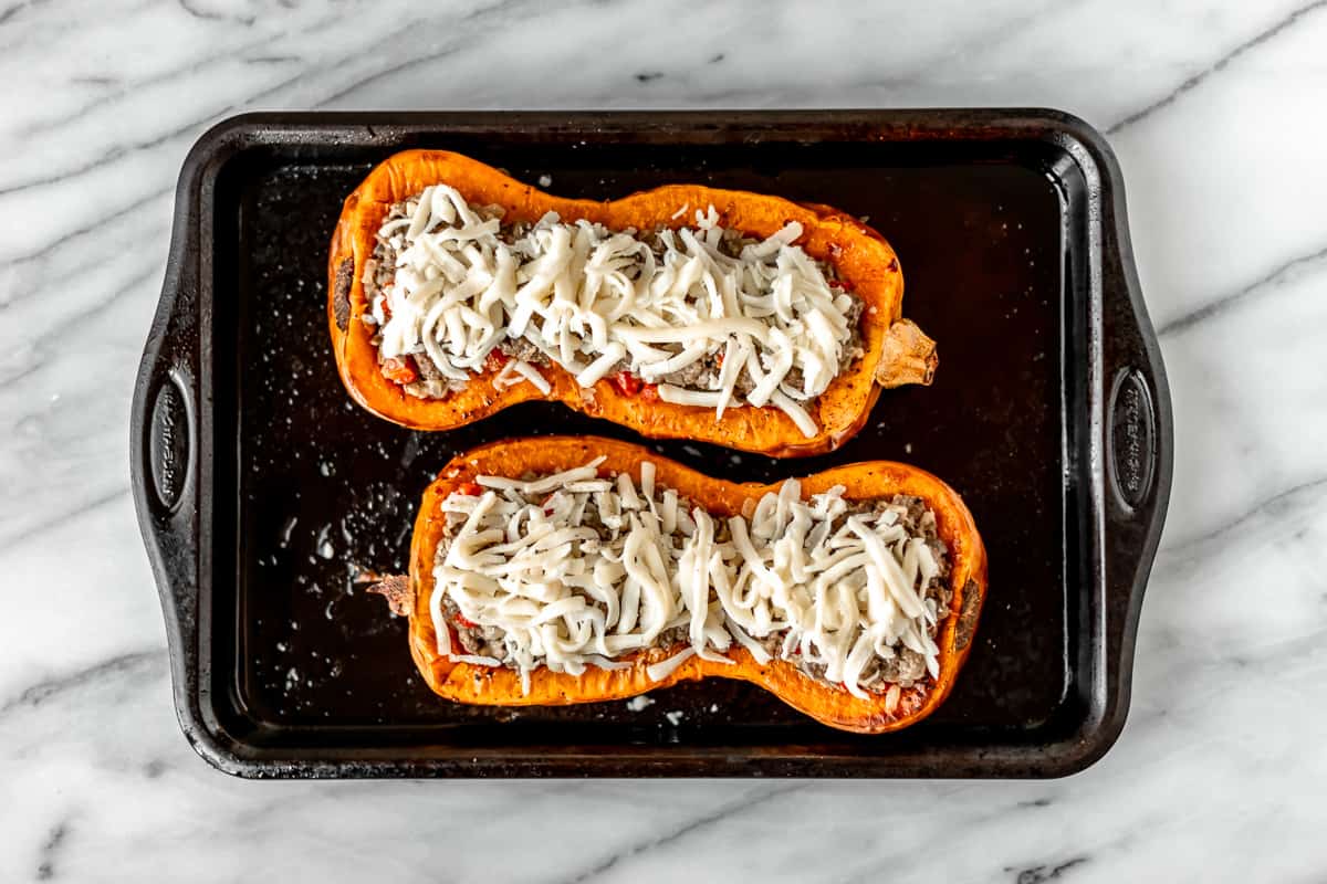 Two butternut squash halves stuffed with a beef mixture and topped with shredded mozzarella cheese on a baking sheet.