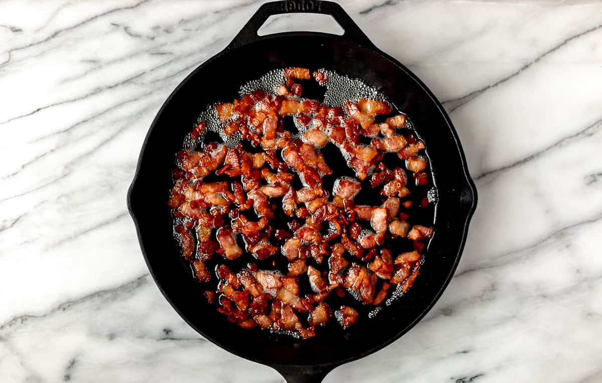 Diced bacon cooking in a cast iron skillet over a marble background.