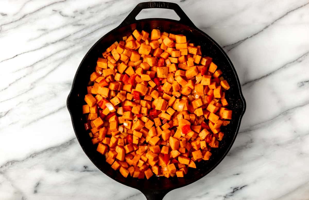 Sweet potatoes, onion and red bell pepper cooking in a cast iron skillet over a marble background.