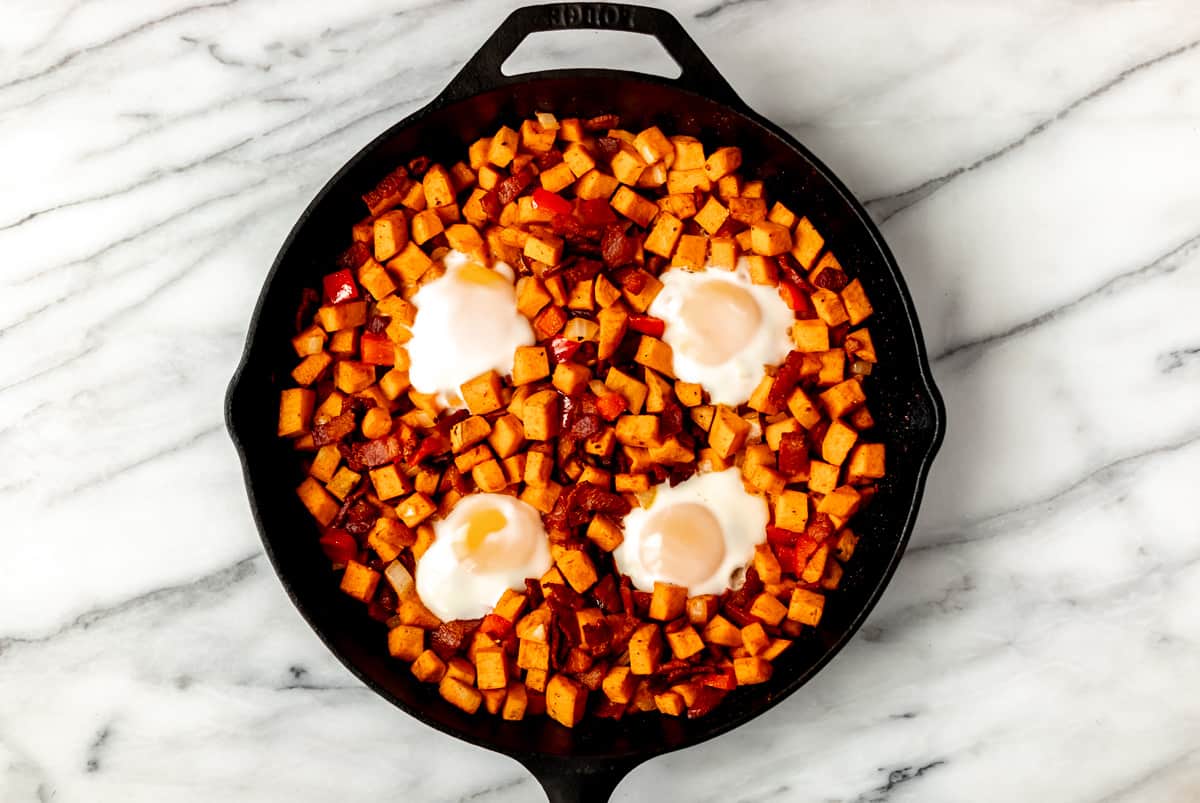 Baked sweet potato hash with 4 eggs in it in a cast iron skillet over a marble background.