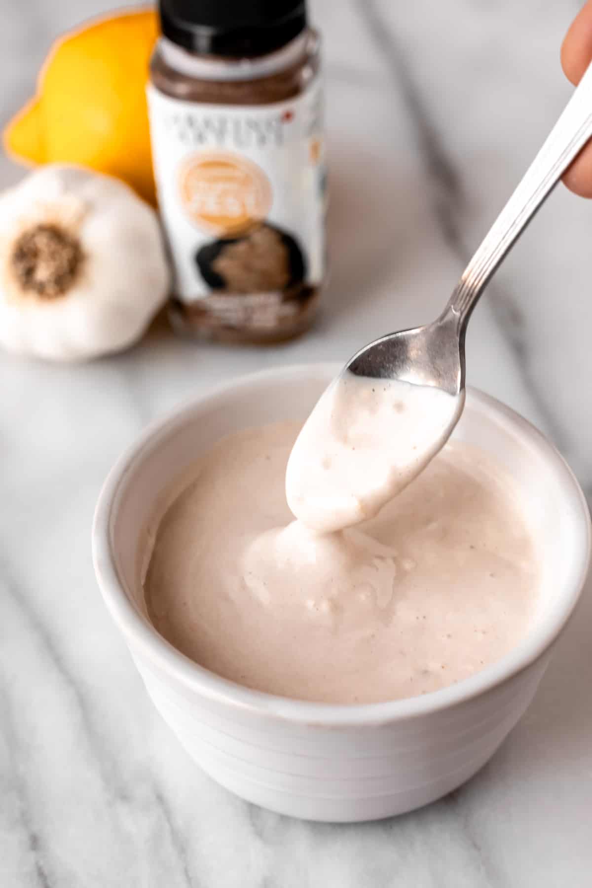 Truffle aioli in a white bowl with a small spoon lifting some up with a lemon, head of garlic, and bottle of truffle powder in the. background.