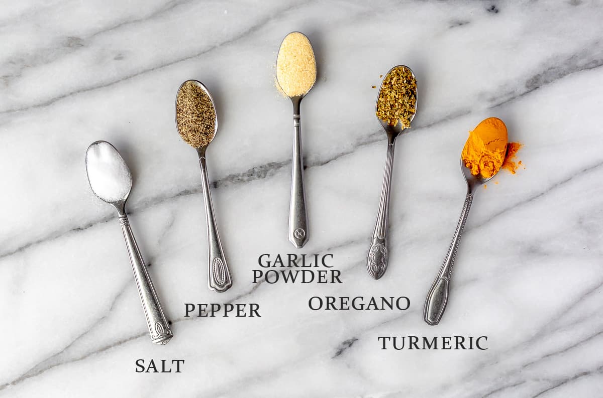 Ingredients needed for homemade adobo seasoning on small spoons on a marble background with text overlay.