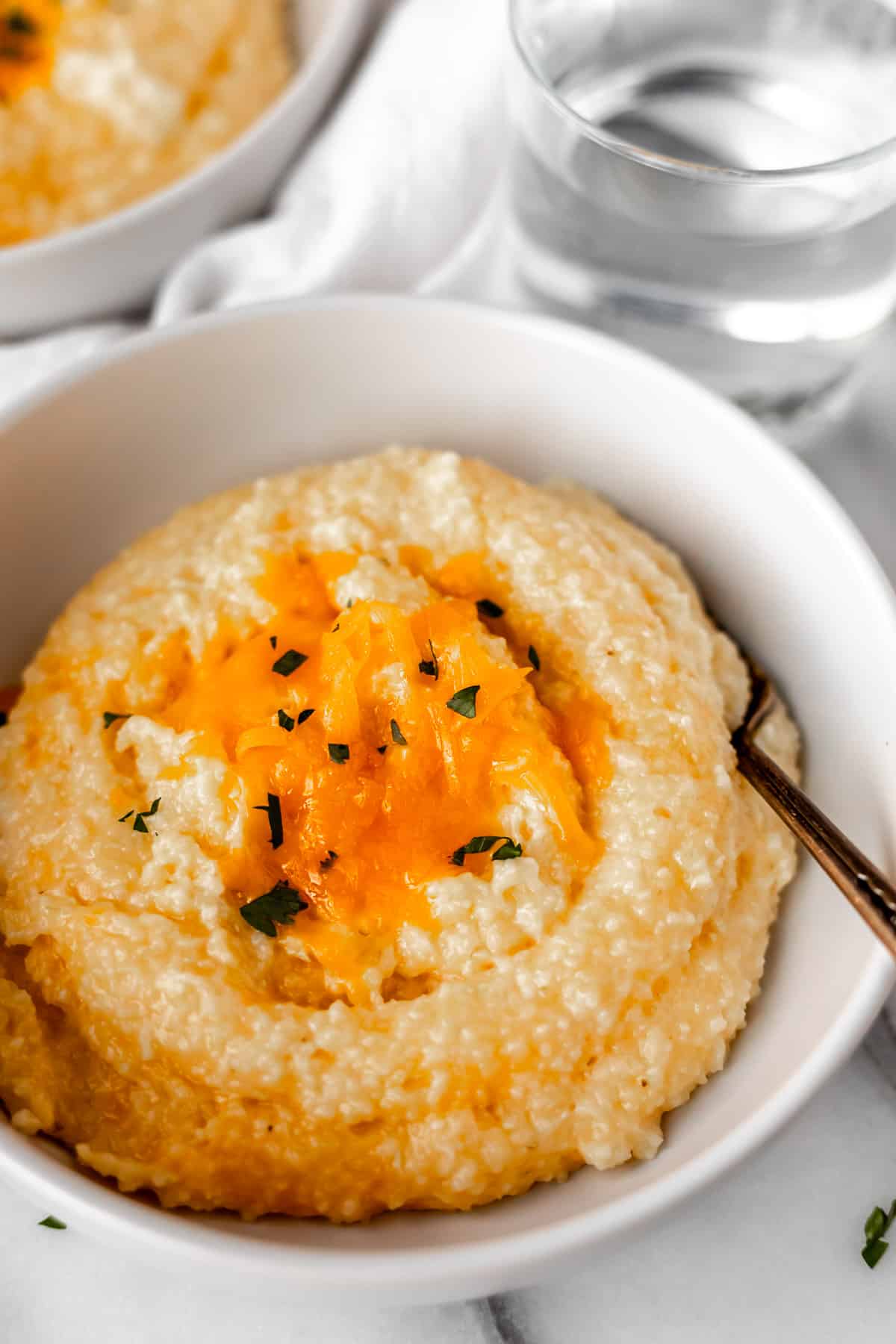 A white bowl of cheddar cheese grits with a fork in it and a second bowl partially showing in the background.