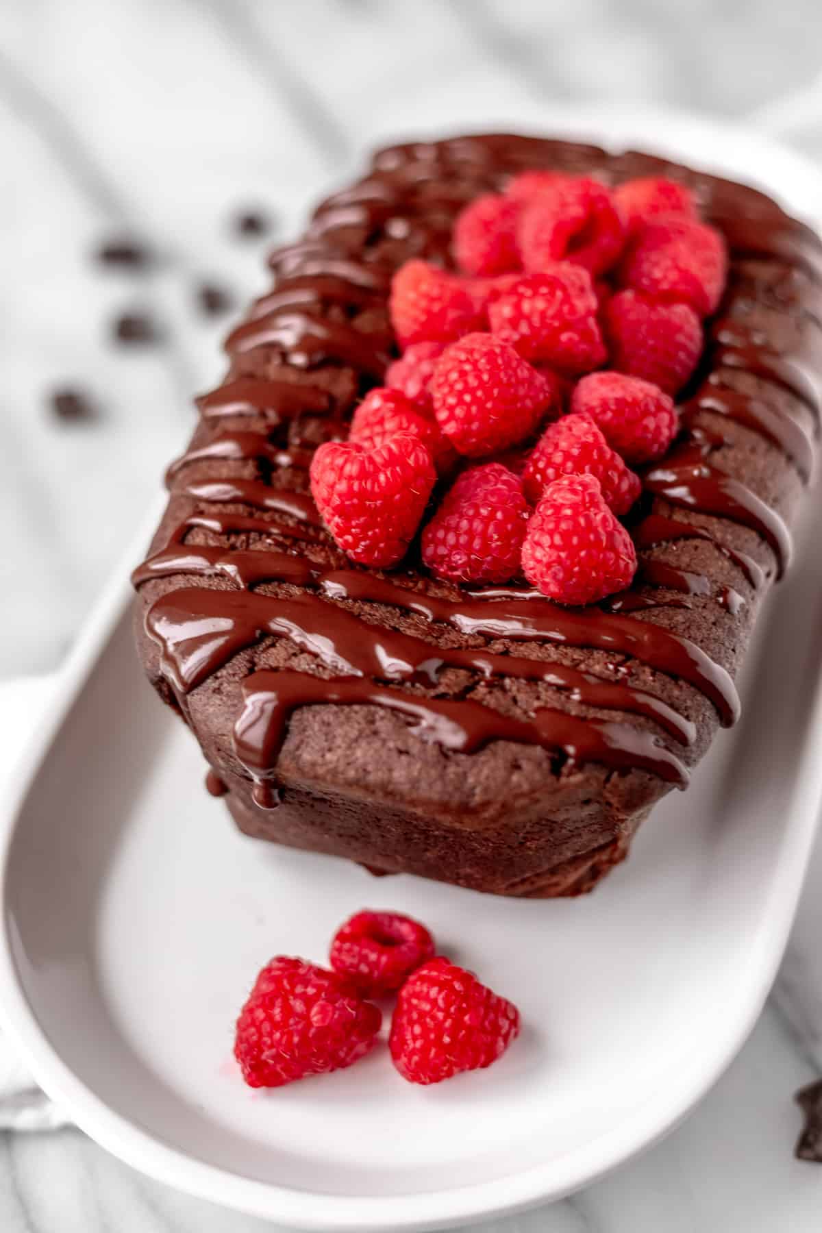 A chocolate pound cake topped with raspberries on a white serving plate.