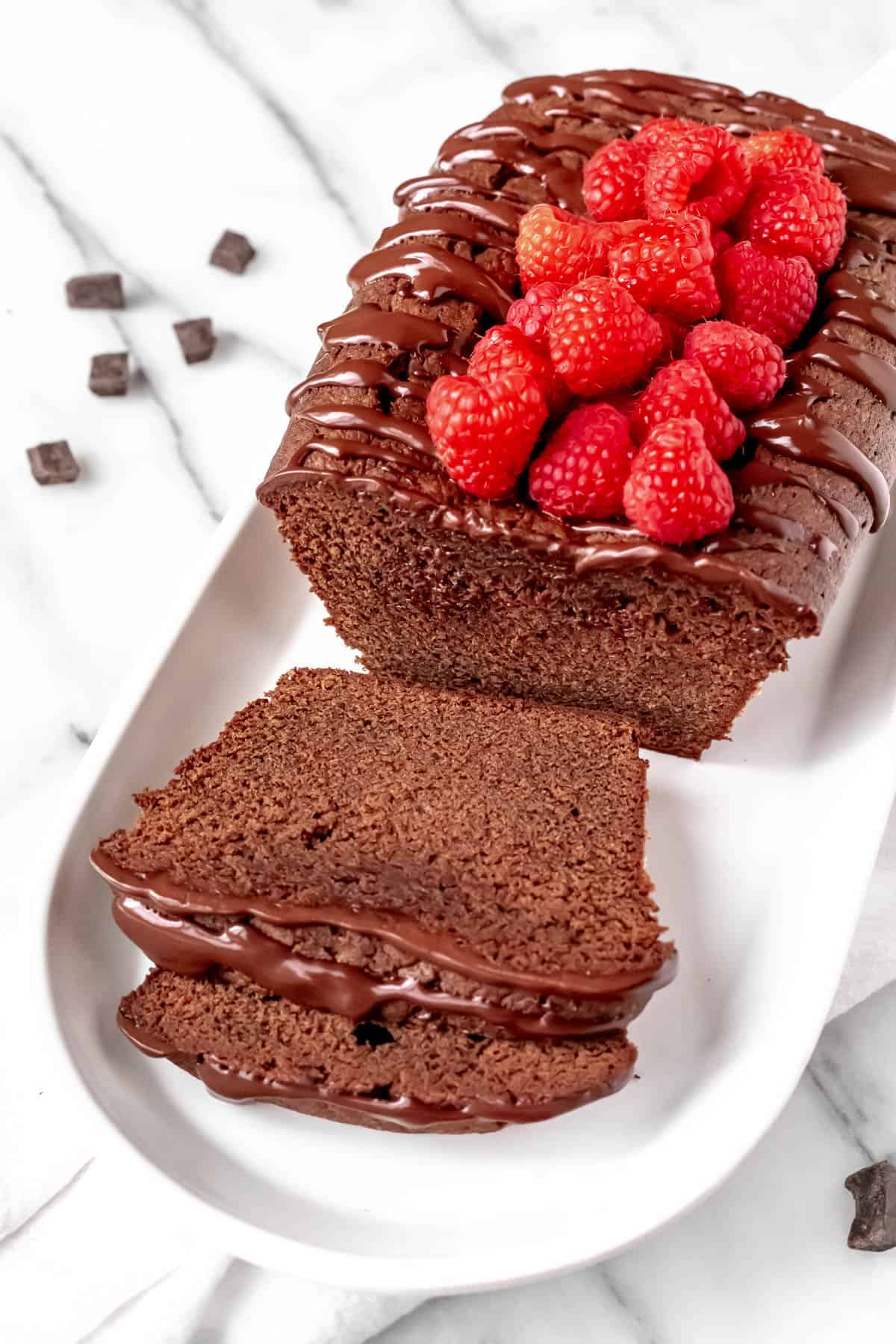 A chocolate pound cake with raspberries on top with 2 slices cut in front of the remaining cake on a white serving plate.