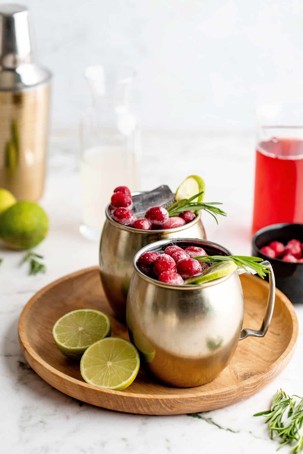 Two Cranberry Moscow Mules in copper mugs on a wood serving tray with limes and other ingredients around it.