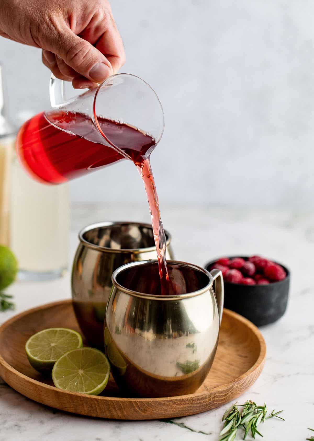 Cranberry juice being poured into a copper mug with a second mug and other ingredients around it.