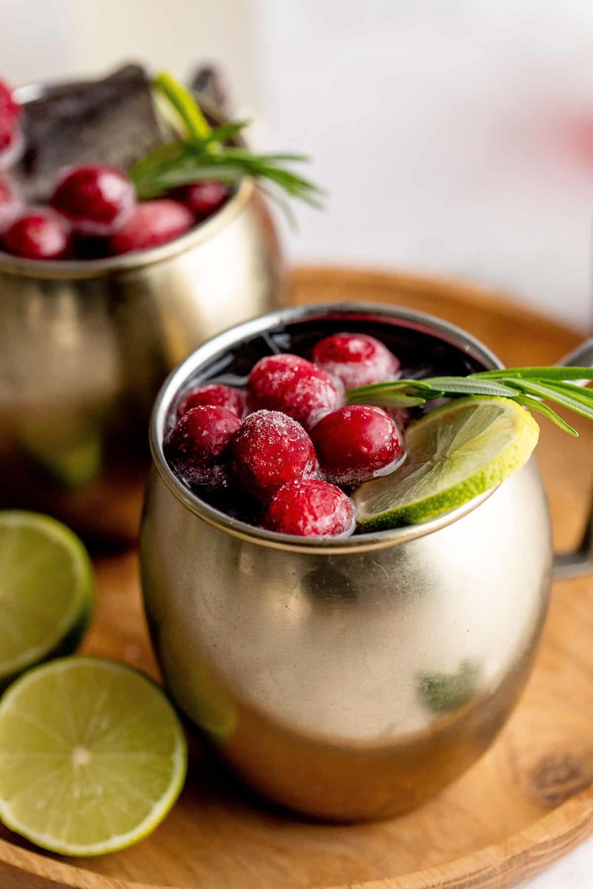A close up of a Cranberry Moscow Mule on a wood serving tray with a second mug and limes around it.