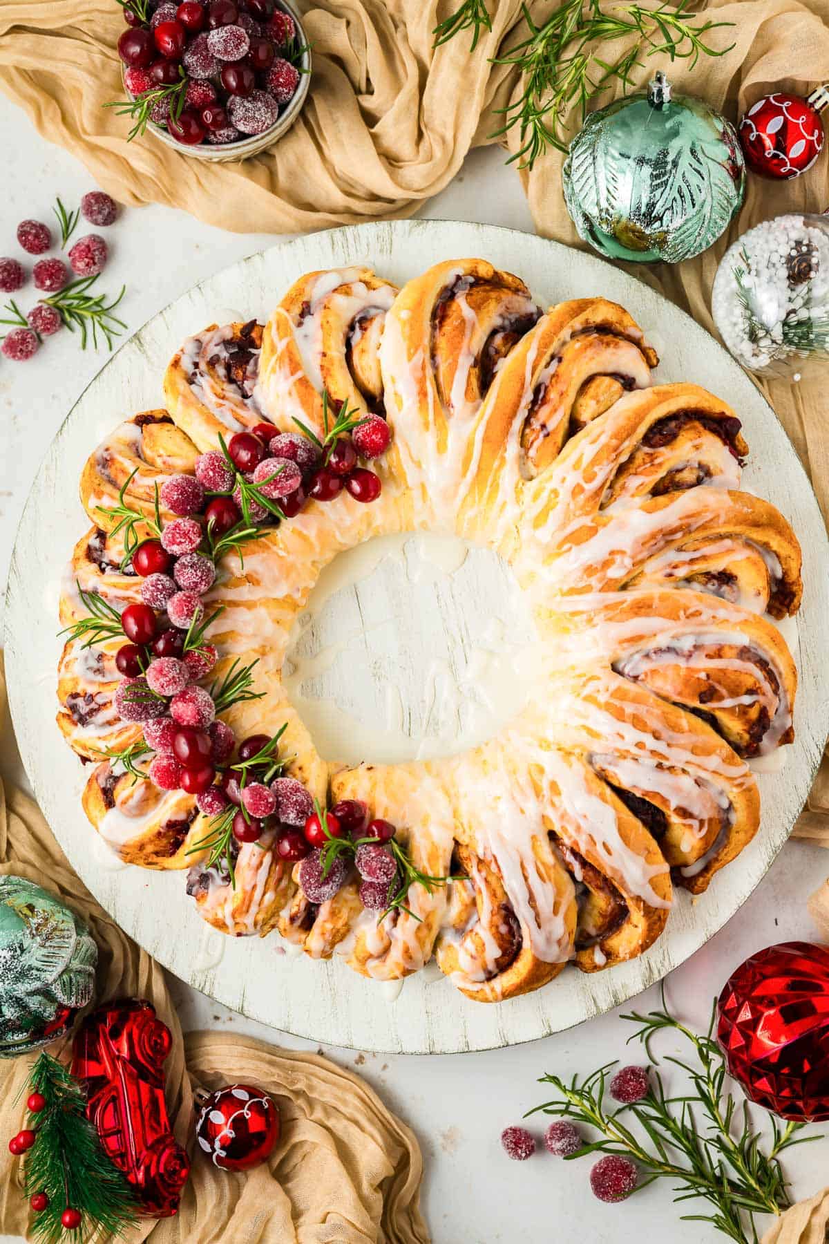 A Cranberry Orange Cinnamon Roll Wreath on a white plate with seasonal ornaments and decor around it.