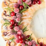 Close up of the top of a cinnamon roll wreath decorated with sugared cranberries and rosemary with text overlay.