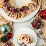 Overhead of a cranberry orange cinnamon roll wreath with a serving on a small plate and ornaments around it with text overlay.