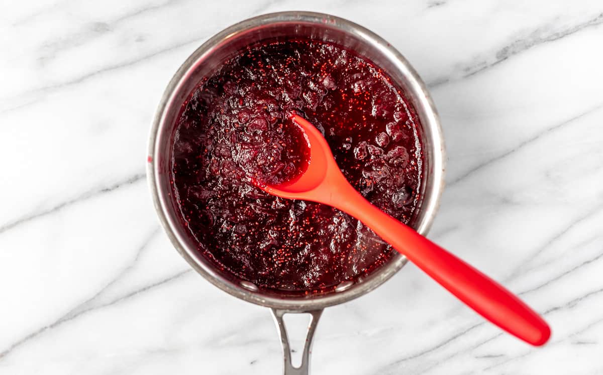 Cooked cranberry sauce in a silver sauce pan with a red spoon in it over a marble background.