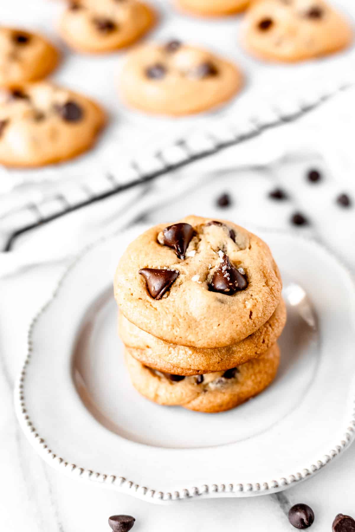 A stack of three dark chocolate chip cookies on a white plate with chocolate chips around it and a tray of more cookies in the background.