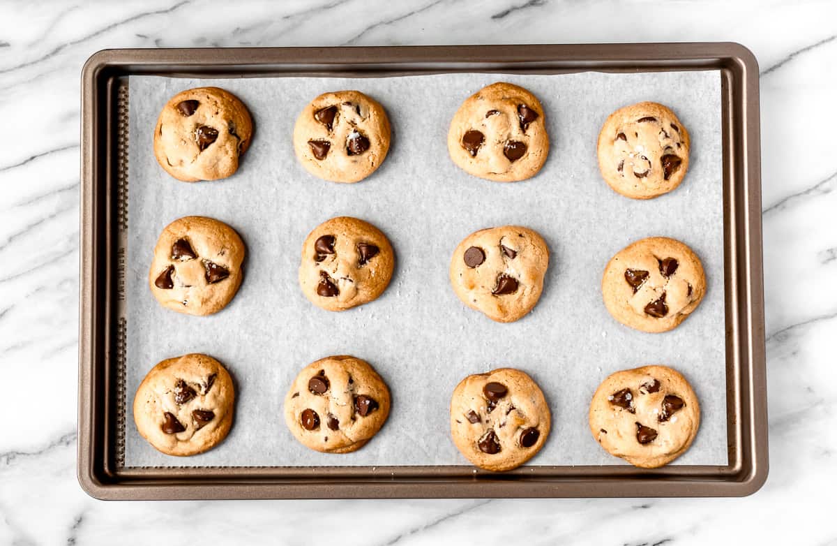 Twelve baked dark chocolate chip cookies on a parchment paper lined baking sheet.