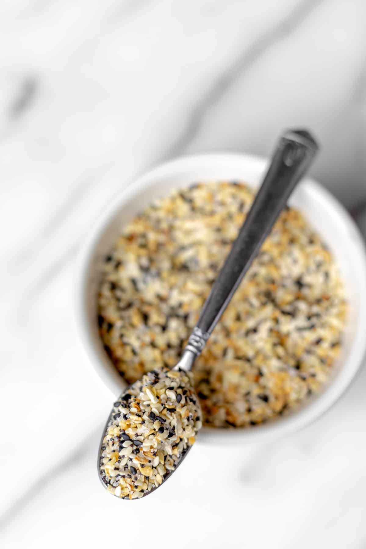 Overhead view of a small white bowl filled with everything bagel seasoning with a spoonful resting on top of the bowl.