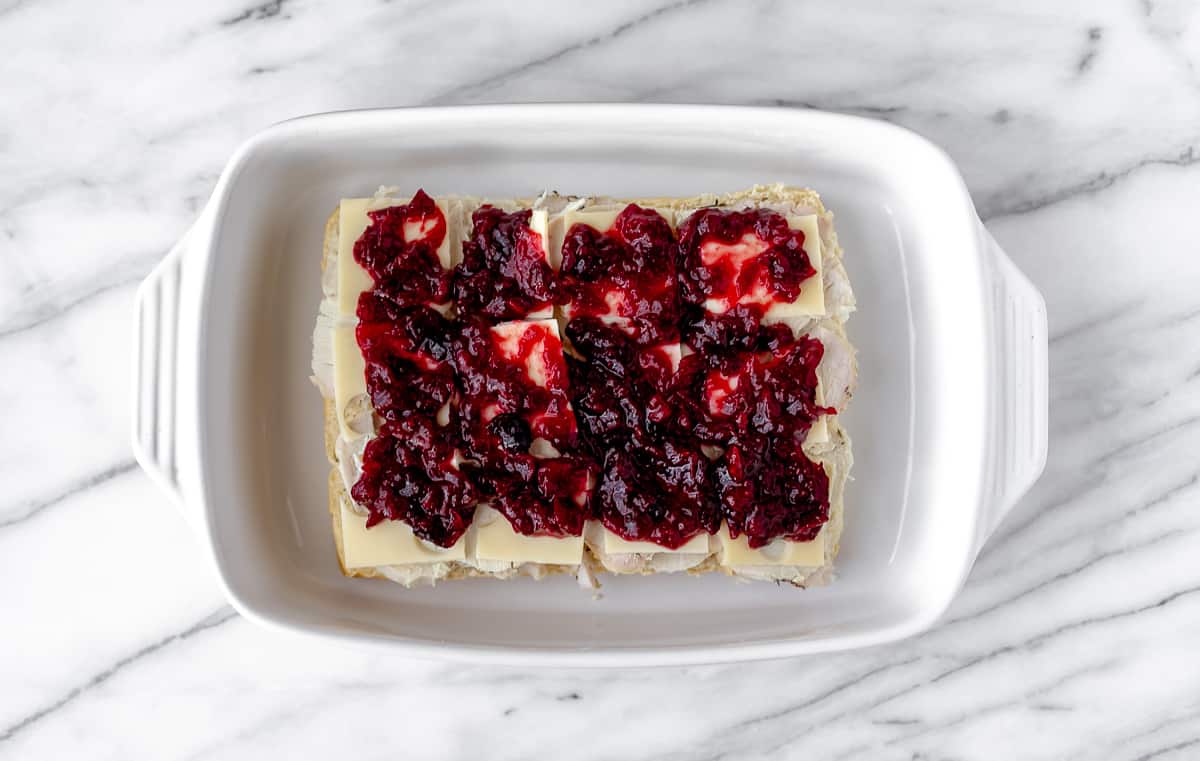 Slices of turkey and Swiss cheese topped with cranberry sauce on the bottom half of Hawaiian Rolls in a white casserole dish.