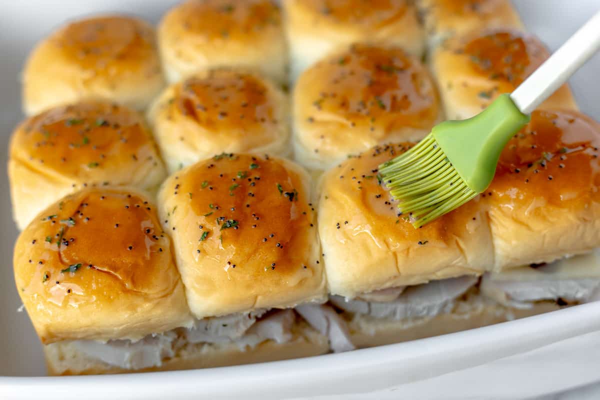 Butter, poppy seeds and parsley being brushed over the tops of Hawaiian rolls.