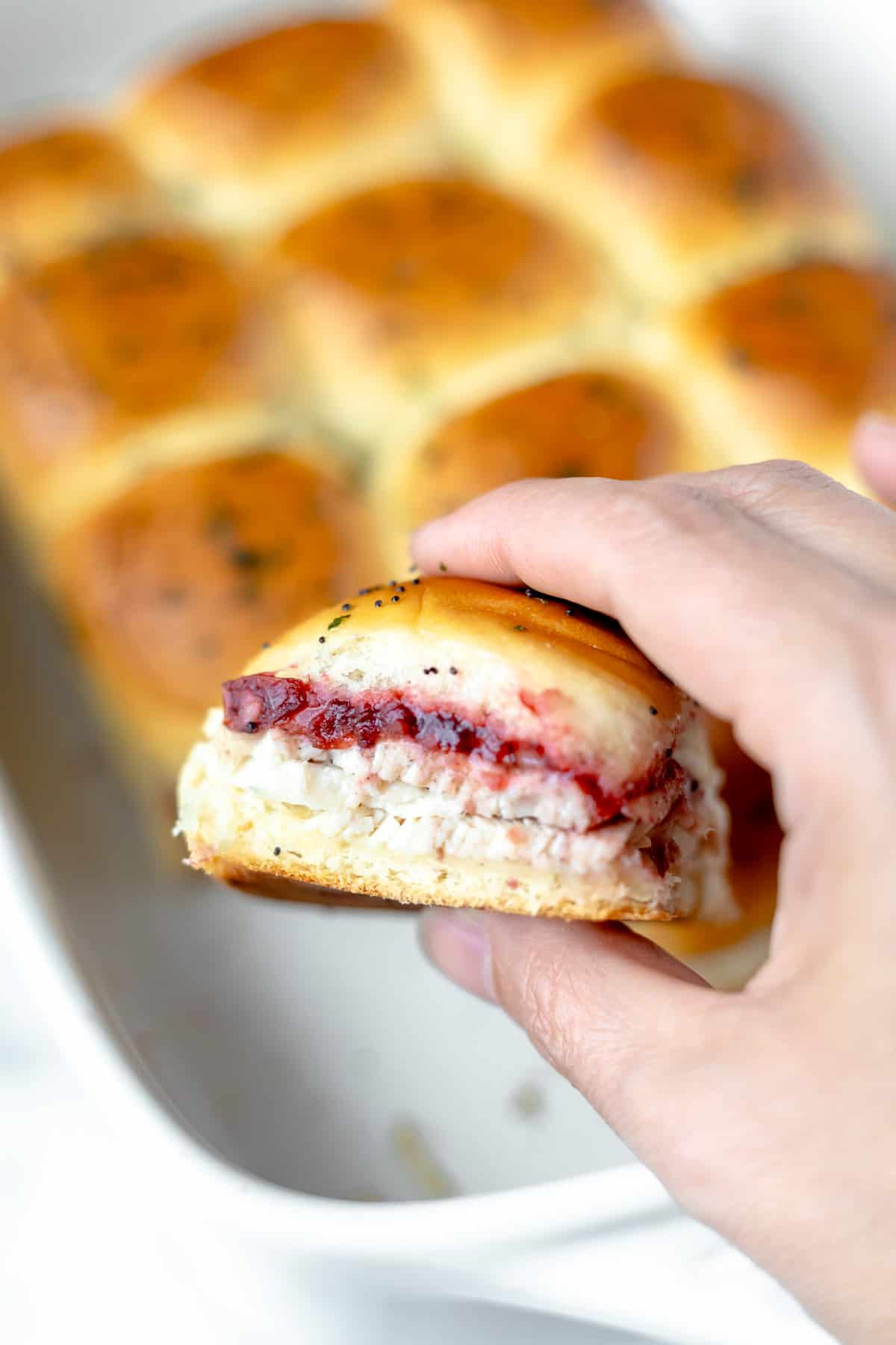 A hand holding a turkey slider in front of a casserole dish of more sliders.