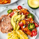 Close up of a plate of chilaquiles rojos topped with slices of jalapeno pepper, tomatoes, and cheese with scrambled egg, refried beans next them and a second plate, avocado, and cherry tomatoes in the background with text overlay.