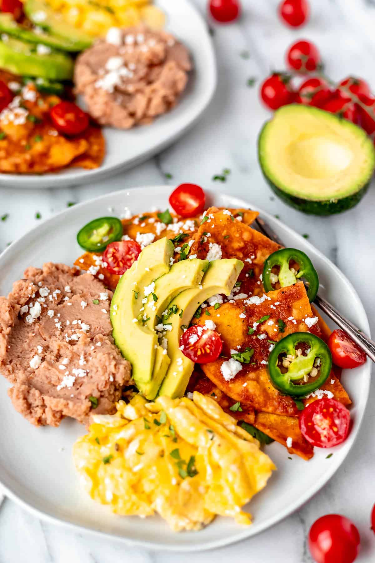 Close up of a plate of chilaquiles rojos topped with slices of jalapeno pepper, tomatoes, and cheese with scrambled egg, refried beans next them and a second plate, avocado, and cherry tomatoes in the background.