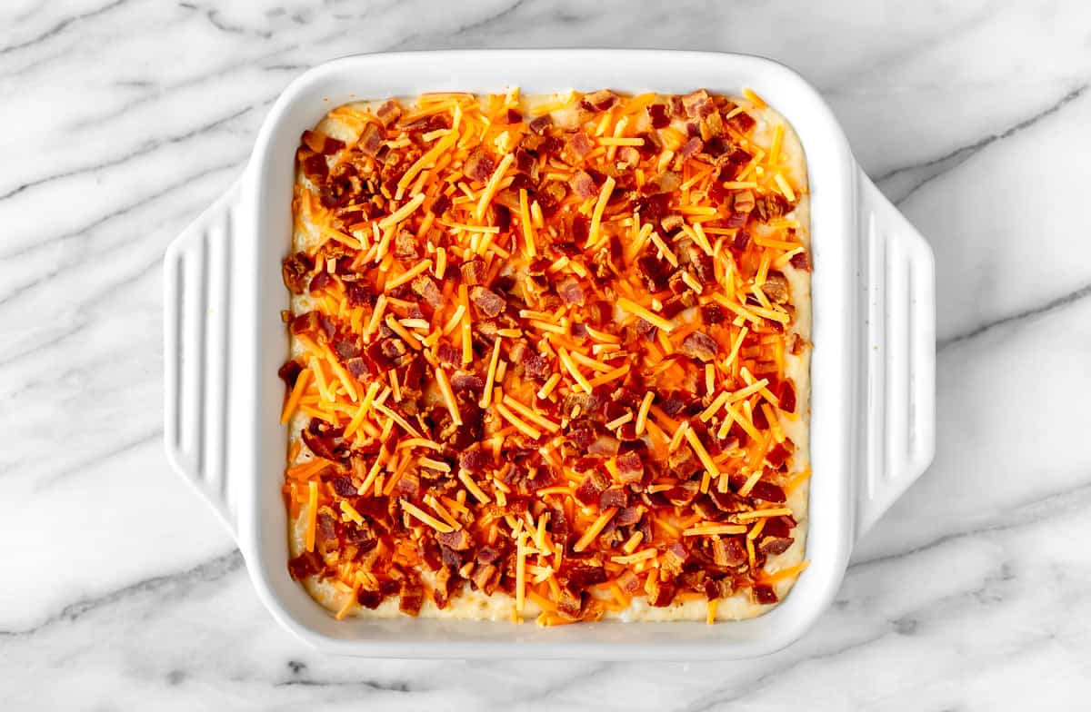 An unbaked grits casserole topped with bacon and shredded cheese.