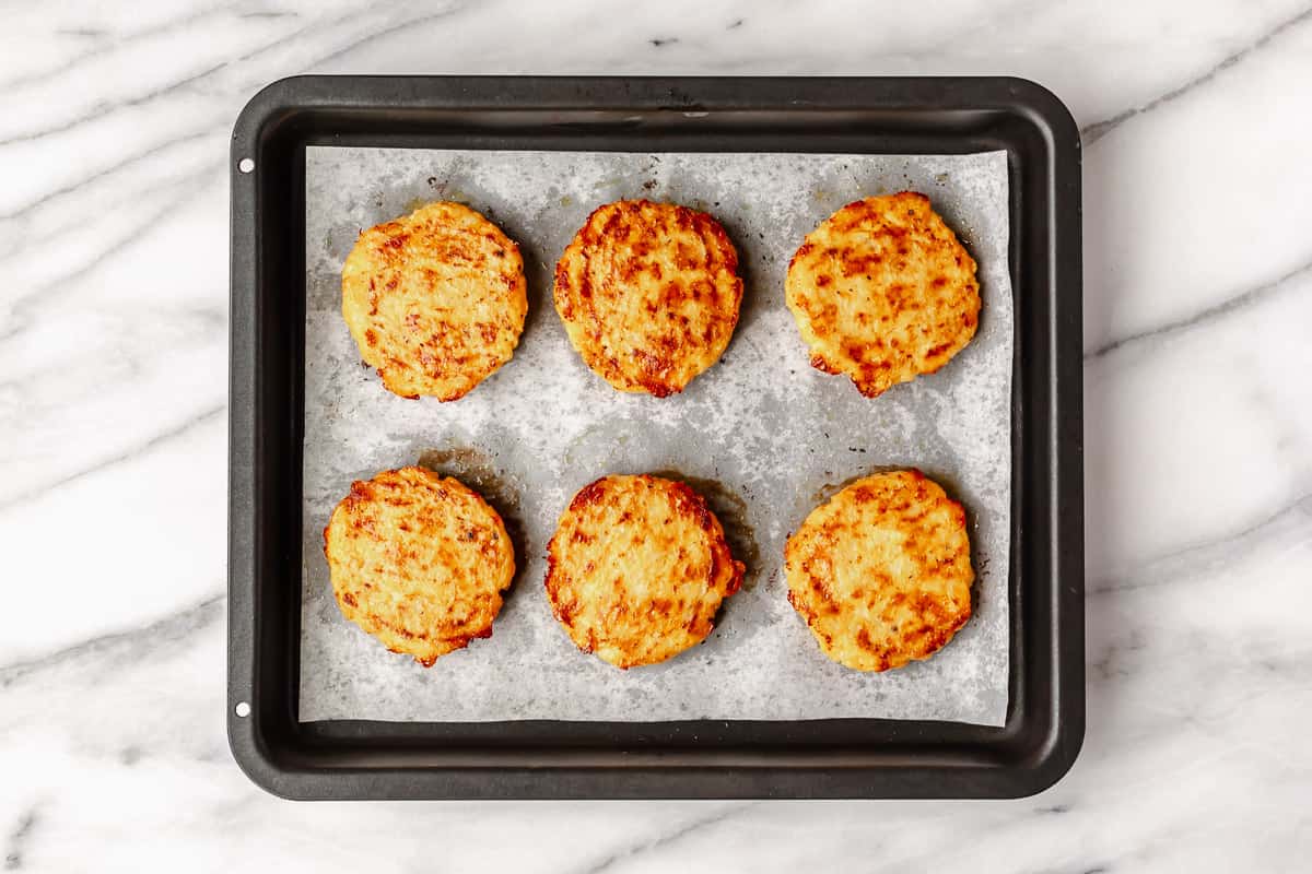 Six baked cauliflower hash browns on a parchment paper lined baking sheet.