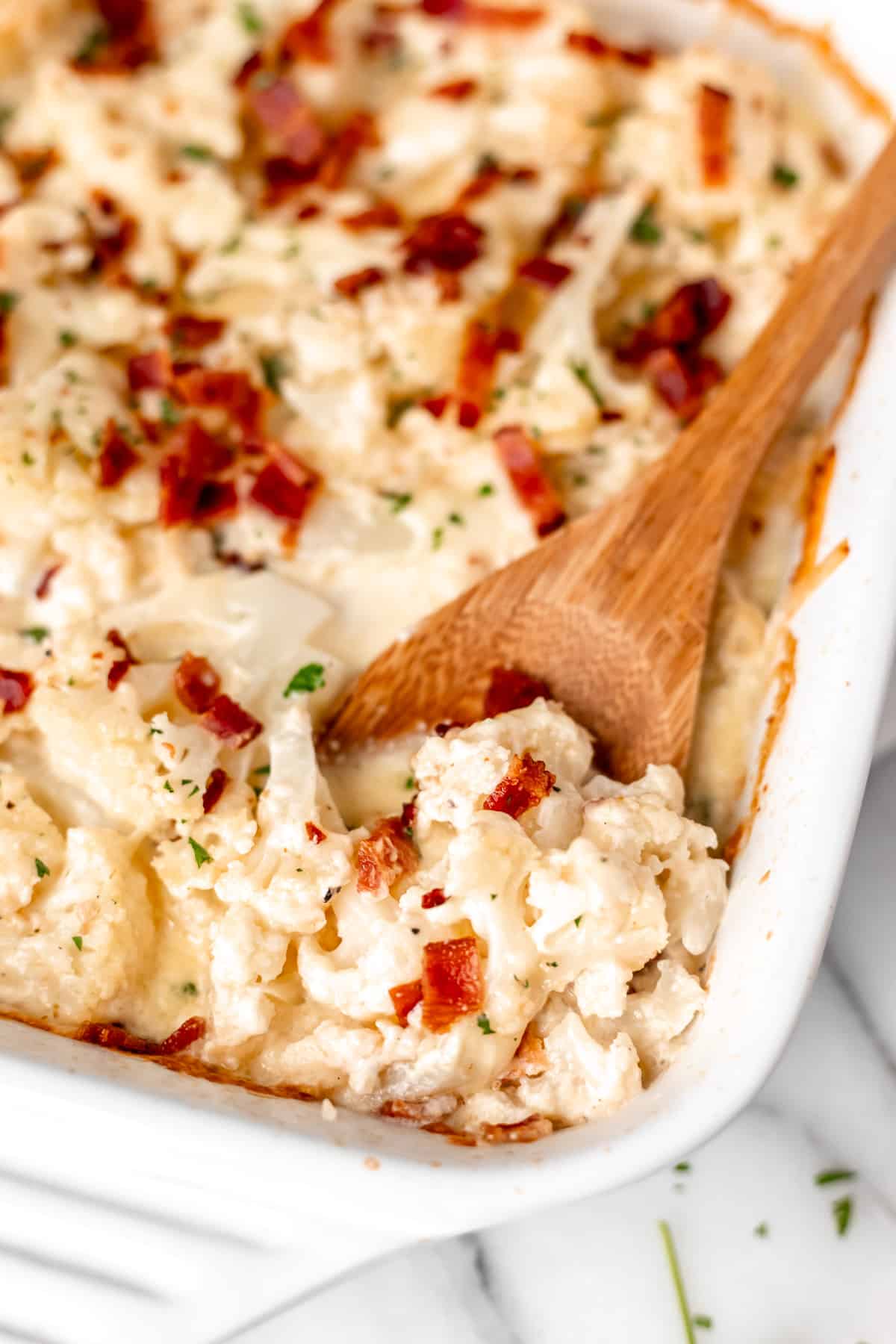 A wood spoon pushing up some keto cauliflower gratin with bacon in a white casserole dish.