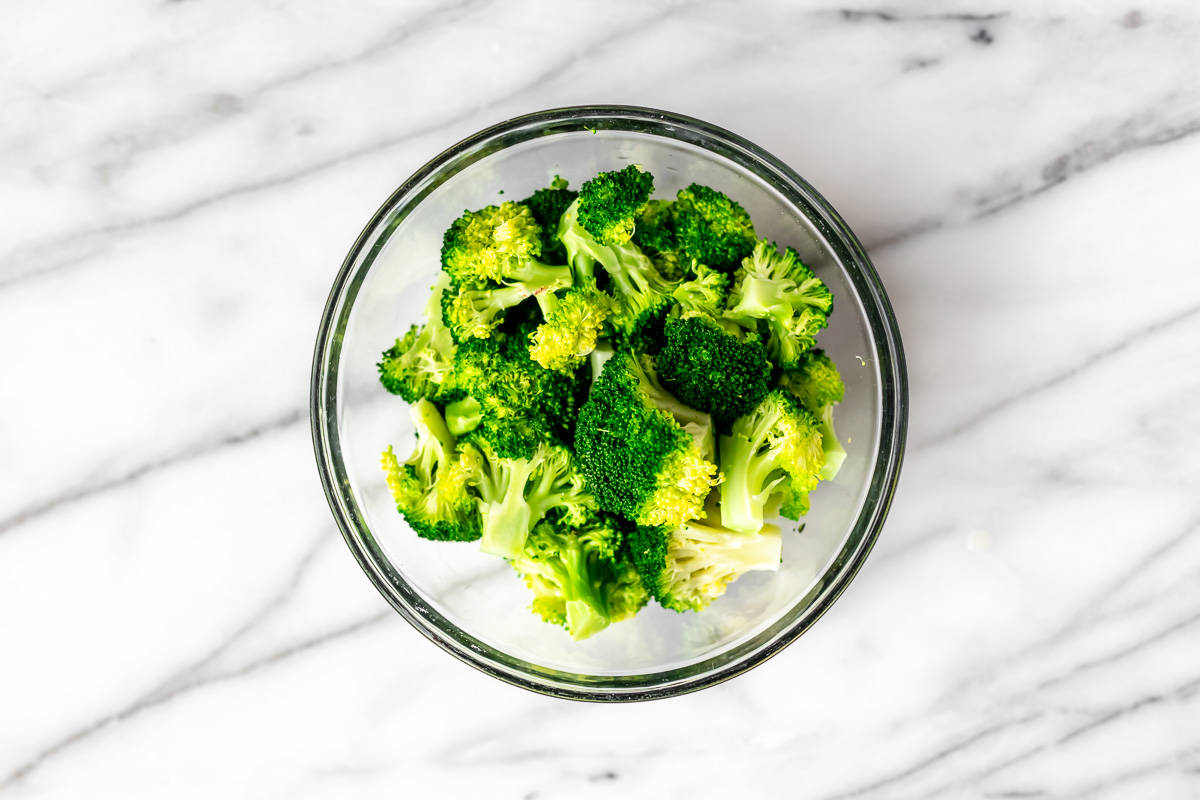 A glass bowl filled with steamed broccoli over a marble background.