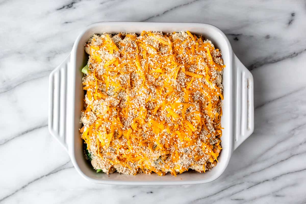 An unbaked keto chicken divan casserole topped with shredded cheddar cheese and bread crumbs.