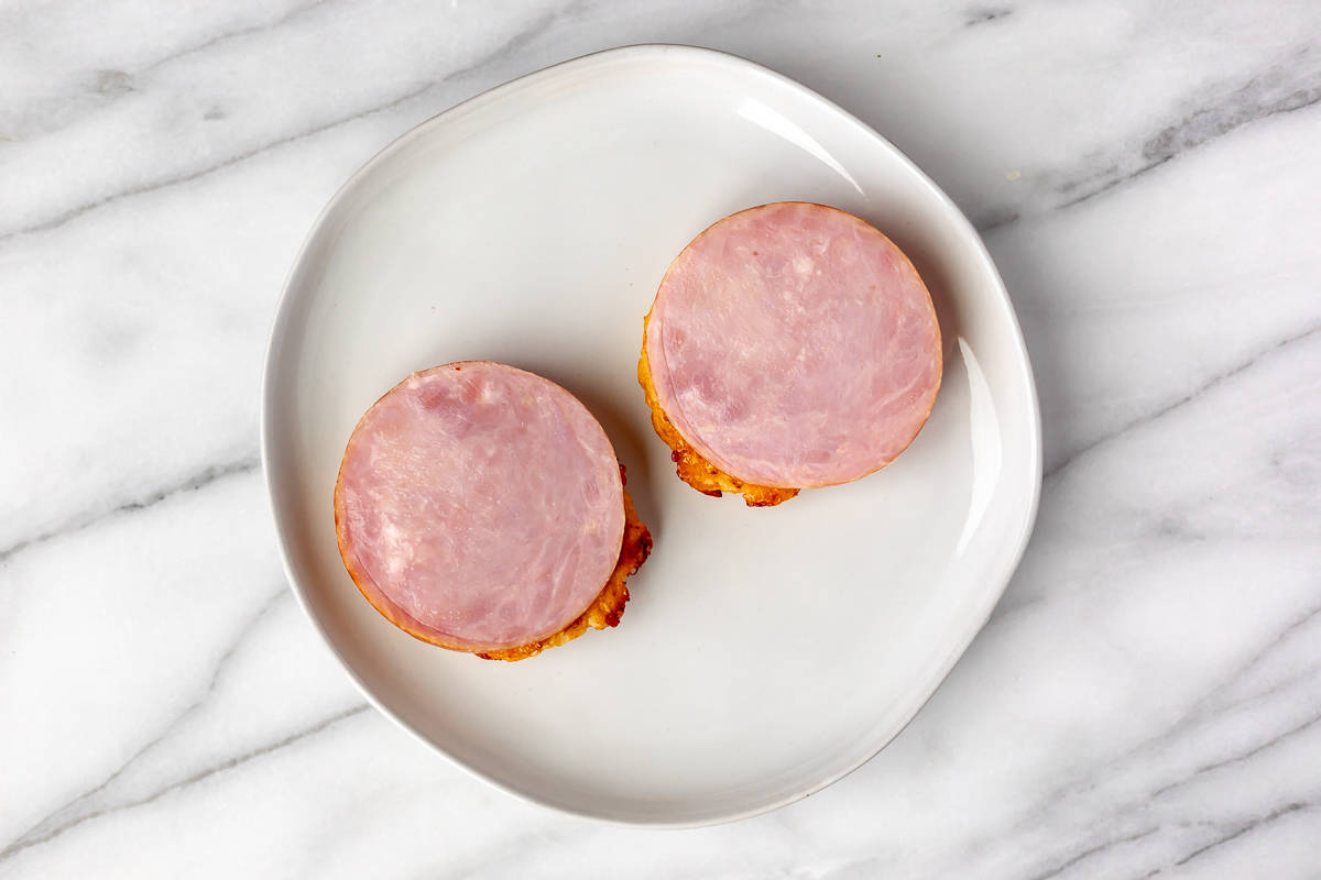 Two cauliflower hash browns topped with a slice of Canadian bacon on a white plate over a marble background.