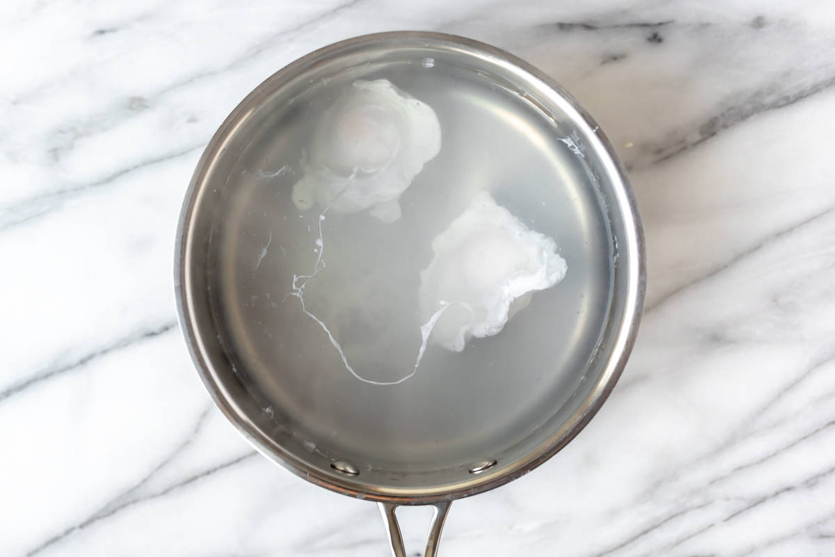 Two poached eggs in a pot of water over a marble background.