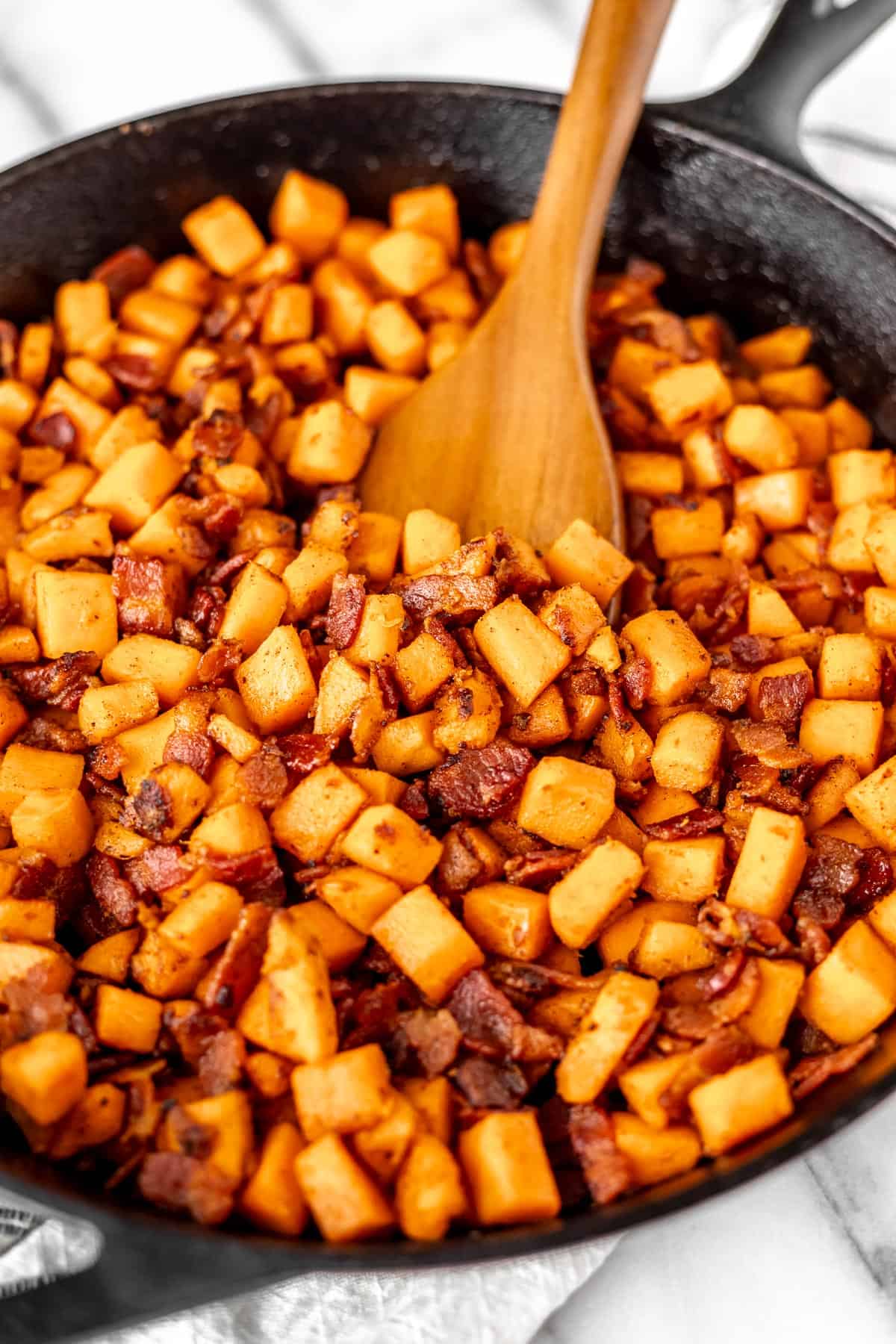 A skillet of maple bacon sweet potato hash with a wood turner pushing some up.