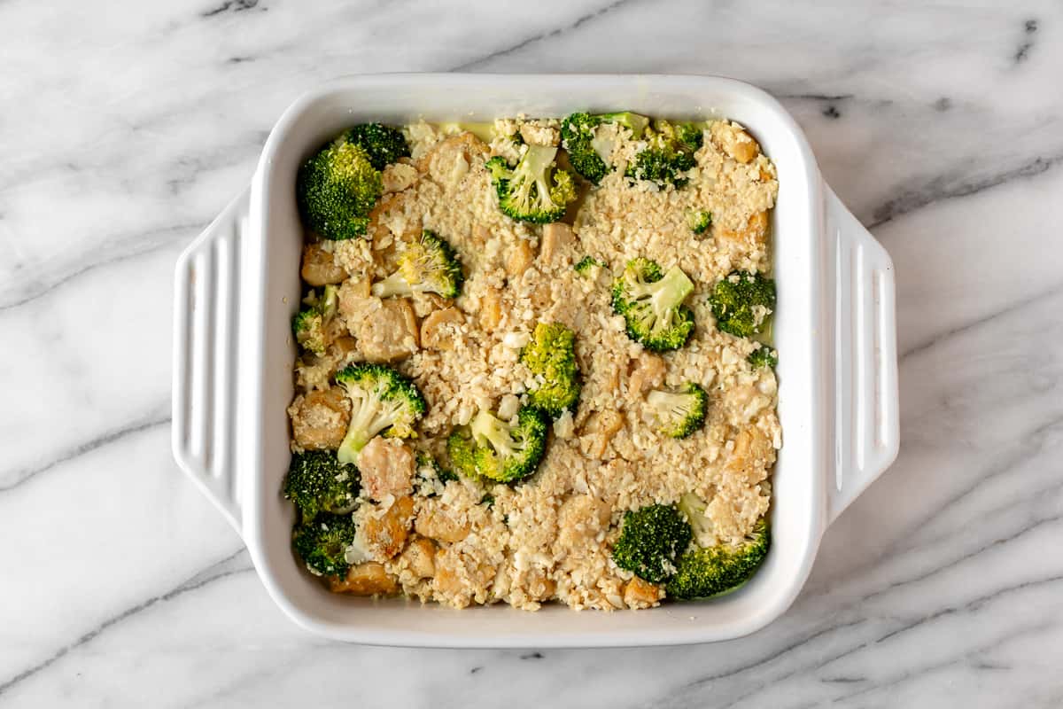 An unbaked chicken and broccoli casserole with cauliflower rice in a white, square casserole dish on a marble background.