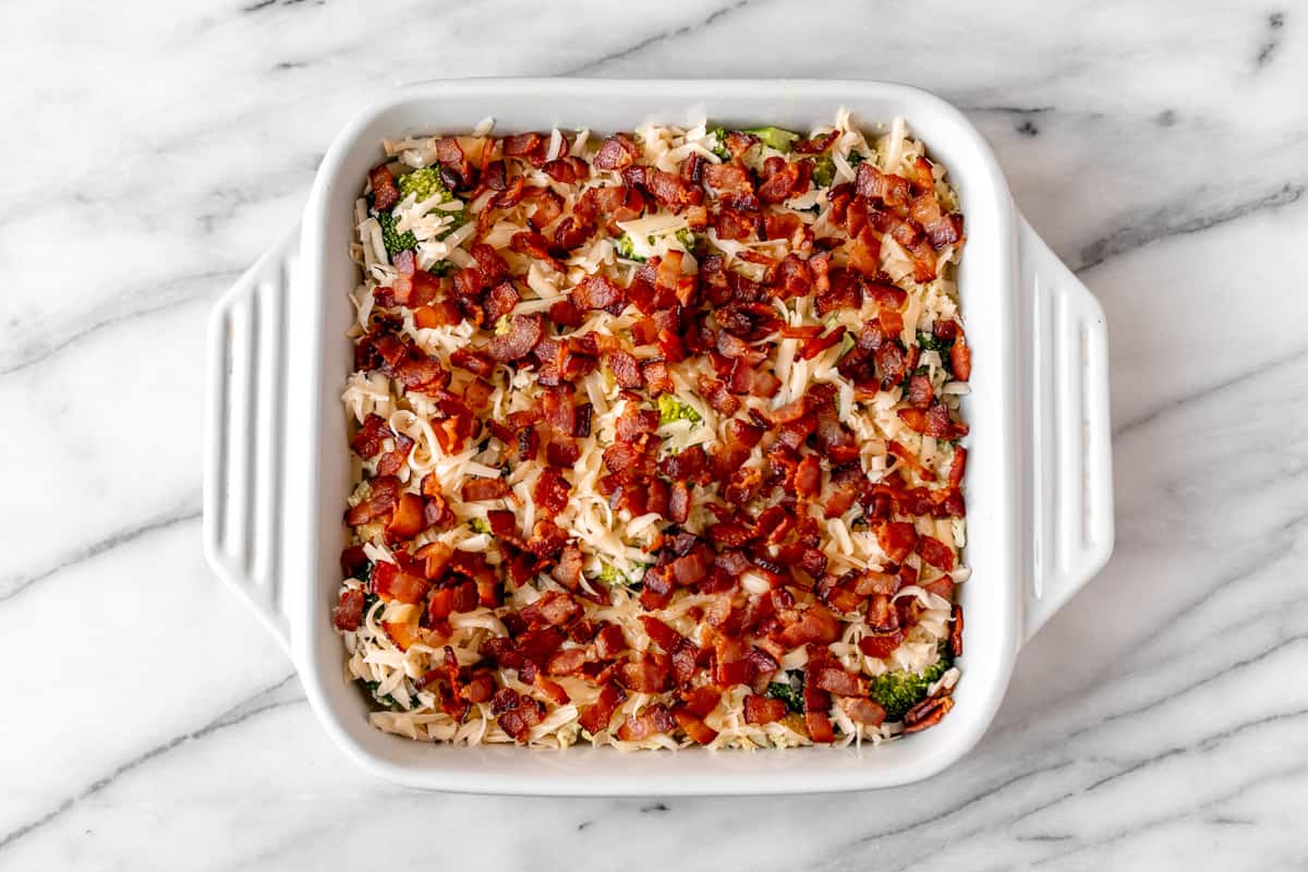 An unbaked chicken and broccoli casserole topped with bacon and cheese in a white, square casserole dish.