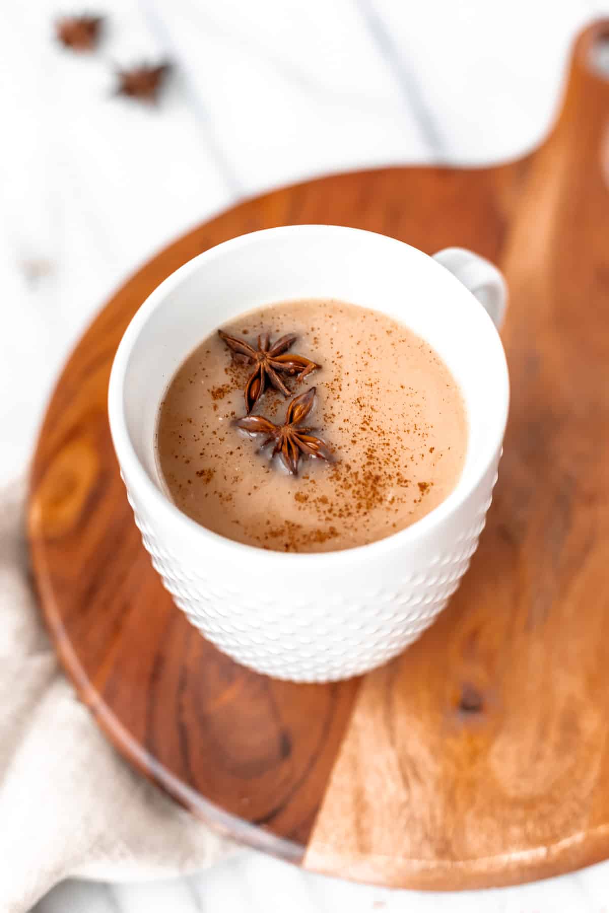 A coconut chai latte in a white mug on a wood board with two star anise and spices on top.
