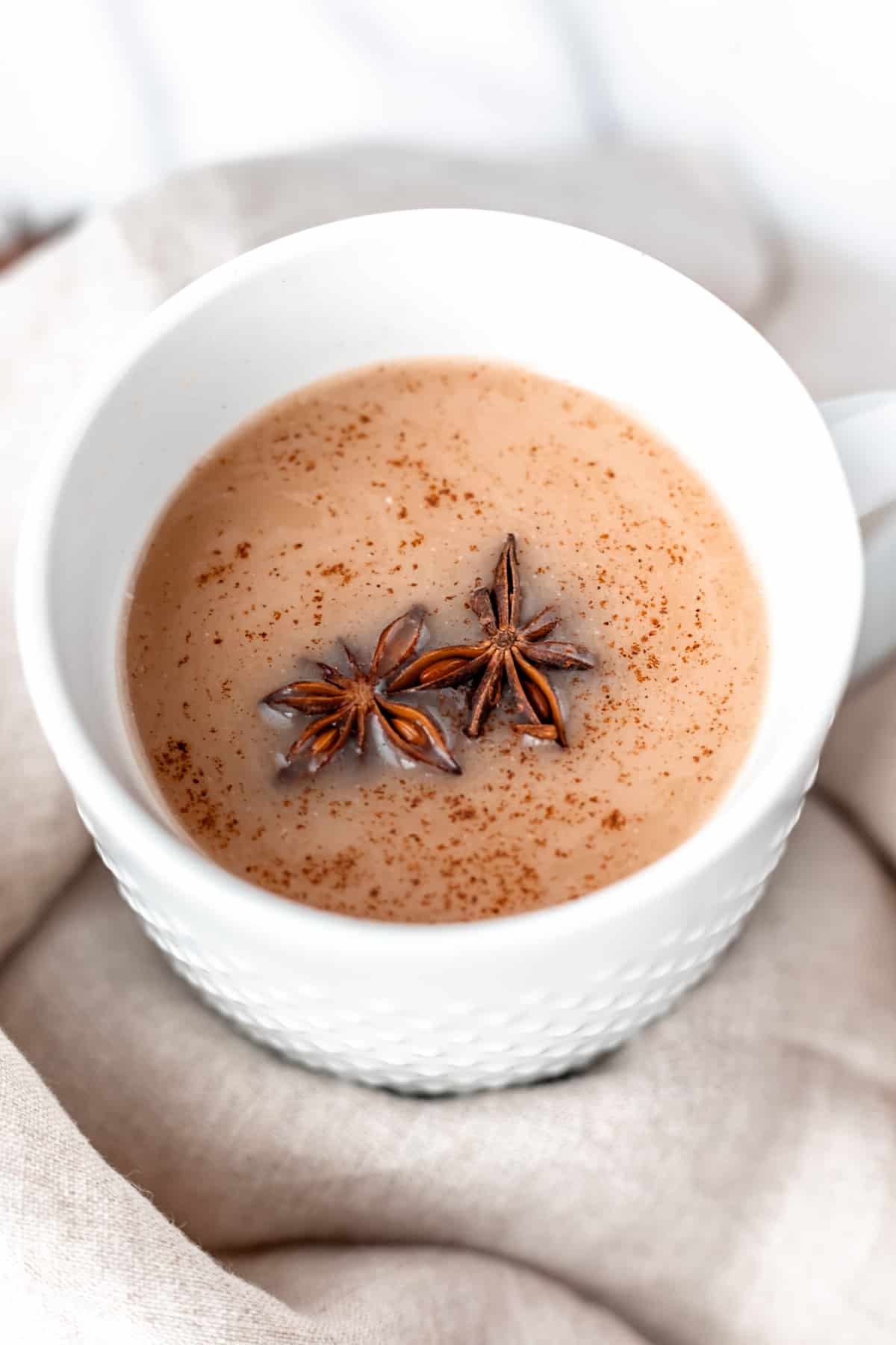 Close up of a coconut chai tea latte in a white mug with two star anise and spices on top with a linen towel around it.